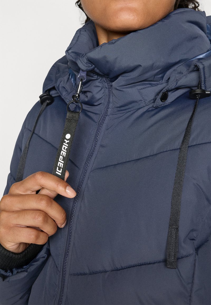 Doudoune marine avec col haut, présentant un extérieur texturé, des cordons de serrage réglables et une étiquette de marque noire près de la fermeture éclair.