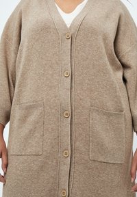 Cardigan beige oversize realizzato in morbido maglione, con scollo a V, cinque bottoni e due tasche frontali con bordi a costine.