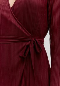 Bordeaux wikkeljurk met verticale ribbels, diepe V-hals, lange mouwen en een gestrikte tailleband. Glad materiaal met een subtiele glans.