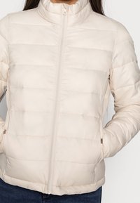 Beige prošívaná bunda s vertikálním prošíváním, vysokým límcem a zipovým uzávěrem vpředu. Hladký povrch se bočními kapsami.