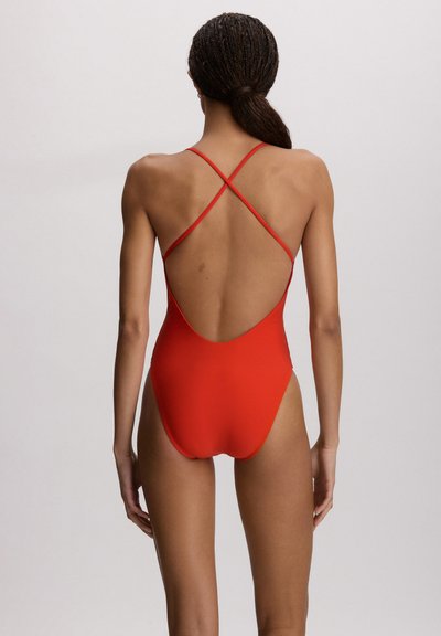 Kobieta z zaplecionymi włosami w czerwonym, jednolitym kostiumie kąpielowym z niskim dekoltem na plecach i cienkimi, krzyżującymi się ramiączkami, stojąca na tle jasnego, gładkiego tła.
