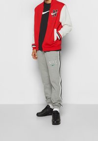 Chaqueta varsity roja y blanca con cremallera negra, camiseta negra, pantalones de chándal grises con el logo "NIKE" y zapatillas negras. Conjunto deportivo casual.
