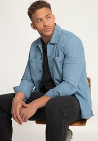 Lichtblauwe denim shirt met een kraag, knoopsluiting aan de voorkant en twee borstzakken, gedragen over een zwart t-shirt, in combinatie met zwarte broek.