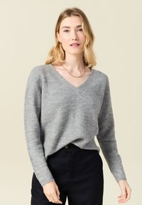 Grauer V-Ausschnitt-Pullover mit weicher Textur, langen Ärmeln und lockerer Passform, kombiniert mit dunklen Hosen. Schlichtes Design, ohne Muster oder Akzente.