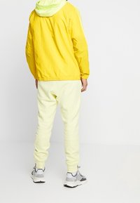 Žlutá lehká bunda s kapucí a zelenou kapucí, v kombinaci se světlými žlutými tepláky a šedobílými teniskami. Zobrazeno ze zadního pohledu.
