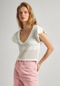 Haut en tricot blanc avec un décolleté en V profond, bord rufflé et motif ajouré, associé à un jean taille haute rose clair.