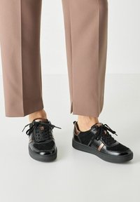 Baskets noires présentant un mélange de matières en cuir verni brillant et en daim mat, avec un accent métallique et des lacets ronds.