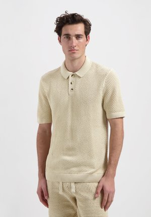 Beige, short-sleeve polo shirt with a textured honeycomb pattern, featuring a ribbed collar, three button placket, and fitted cuffs.