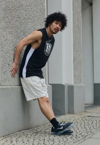 Top sin mangas negro con un logo, panel lateral blanco; pantalones cortos beige; zapatillas deportivas negras con suelas transparentes; de pie contra una pared.