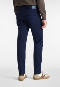 Pantaloni in cotone blu navy con vestibilità dritta, tasche posteriori e una patch in pelle in vita. Indossati con sneakers di colore chiaro.