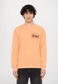 Langærmet t-shirt i lys orange bomuld med et sort "Aries" logo på venstre brystlomme. Simpelt design med ribbede manchetter.