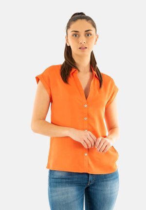 Blusa arancione a maniche corte con colletto, bottoni sul davanti e un tessuto fluente. Indossata con jeans blu.