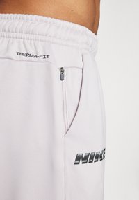 Svetlo šedé nohavice Nike Therma-FIT s elastickým pásom, s bočnou zipsovou vreckom a čiernym logom na stehne. Tkanina s hladkou textúrou.