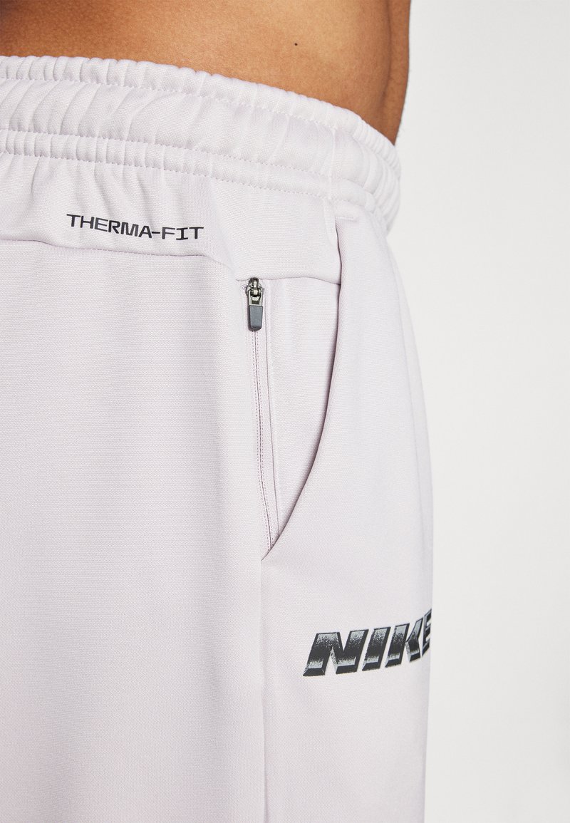Pantalon Nike Therma-FIT gris clair avec une taille élastique, doté d'une poche zippée sur le côté et d'un logo noir sur la cuisse. Tissu à texture lisse.