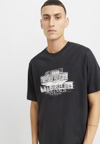 Camiseta de algodón negra con un estampado gráfico blanco de un automóvil y la palabra "ORIGINALS" junto con texto adicional, cuello redondo y mangas cortas.
