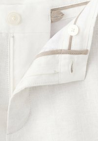 Gros plan de la ceinture d'un pantalon en lin blanc avec un bouton et un bord beige, montrant la boutonnière et la texture du tissu.