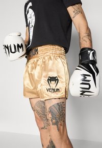 Goldene Satinschlaghosen mit schwarzem "Venum"-Logo und Akzenten, kombiniert mit weißen und schwarzen Boxhandschuhen. Ausführliches Tattoo auf dem Arm sichtbar.