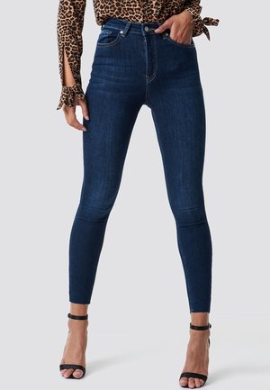 Femme portant un jean skinny bleu marine avec ourlet brut, associé à des sandales à talons hauts noires à bride cheville et un haut à manches longues imprimé léopard.