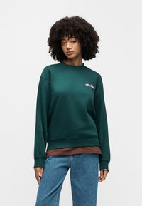 Γυναίκα με σγουρά μαλλιά που φοράει σκούρο πράσινο φούτερ πάνω από καφέ μπλούζα και μπλε τζιν, όρθια μπροστά σε απλό λευκό φόντο.