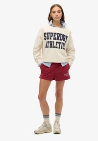Sudadera crema con el texto en azul "SUPERDRY ATHLETIC", superpuesta sobre una camisa de rayas azules. Pantalones cortos burdeos y zapatillas crema con detalles grises.