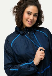 Marineblå lätt jacka med huva, prydd med blå accentlinjer och dragkedja fram. Tyget har en subtil lyster och textur.