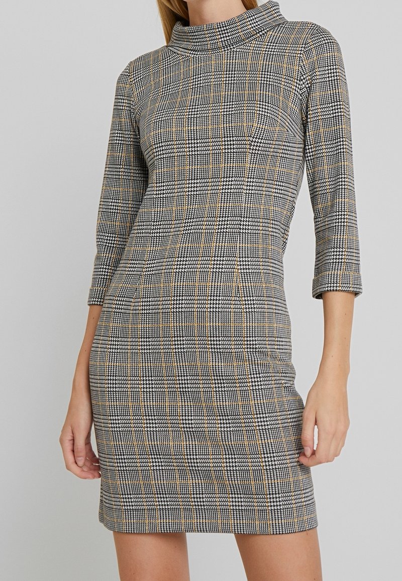 Robe à carreaux en noir, blanc et moutarde, avec un col montant et des manches trois-quarts, confectionnée dans un tissu ajusté et extensible.