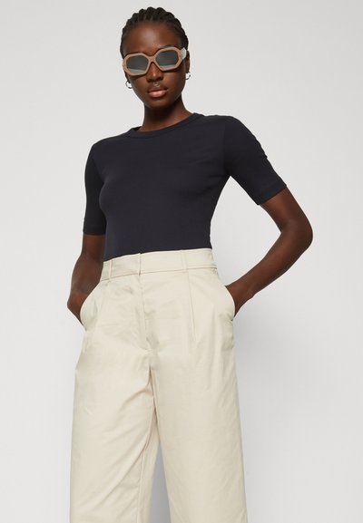 Zwarte geribbelde top met korte mouwen, gecombineerd met hoge taille, crèmekleurige wijde broek. Model draagt oversized getinte zonnebril.