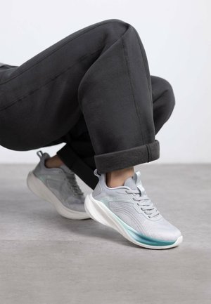 Zapatillas deportivas de color gris claro con una parte superior de malla texturizada, suela blanca suave y un sutil acento turquesa; combinadas con pantalones de chándal negros enrollados.