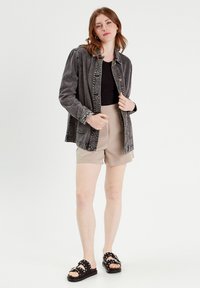 Giacca in denim grigio chiaro, leggera e sbiadita, con chiusura a bottoni, abbinata a una maglietta nera e pantaloni corti beige. Sandali neri con borchie bianche.
