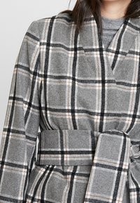 Manteau en laine gris avec un motif à carreaux comprenant des lignes noires, blanches et brunes. Il a une taille ceinturée et un col à revers.