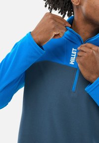 Blau-grauer Halbzip-Pullover aus strukturiertem Stoff, mit erhöhtem Kragen, Millet-Logo auf der Brust und Kontrastnähten.