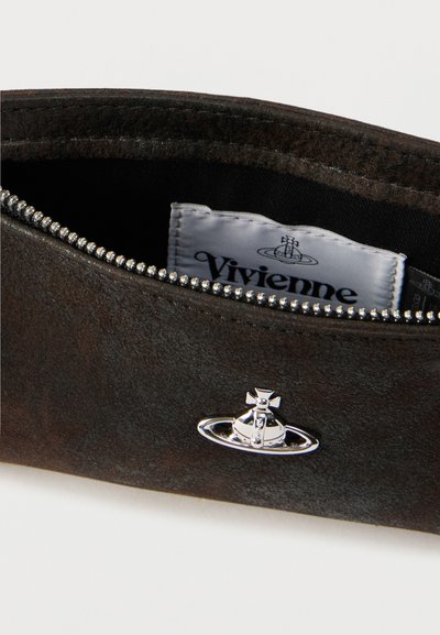 Κοντινό πλάνο σκούρου σουέτ θήκης με ασημένιο φερμουάρ και λογότυπο της Vivienne Westwood σε σχήμα σφαίρας μπροστά, που δείχνει την λευκή ετικέτα της μάρκας μέσα.