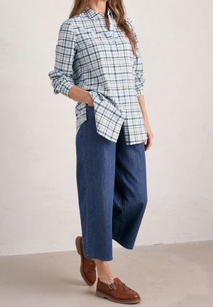 Femme portant une chemise à manches longues à carreaux bleus et blancs, un jean large bleu et des mocassins en cuir marron, debout avec une main dans la poche.