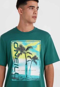 Camiseta de algodón verde con un gráfico de palmera en aqua y amarillo, con el texto "PRIMER NOMBRE EN EL AGUA" debajo del diseño.