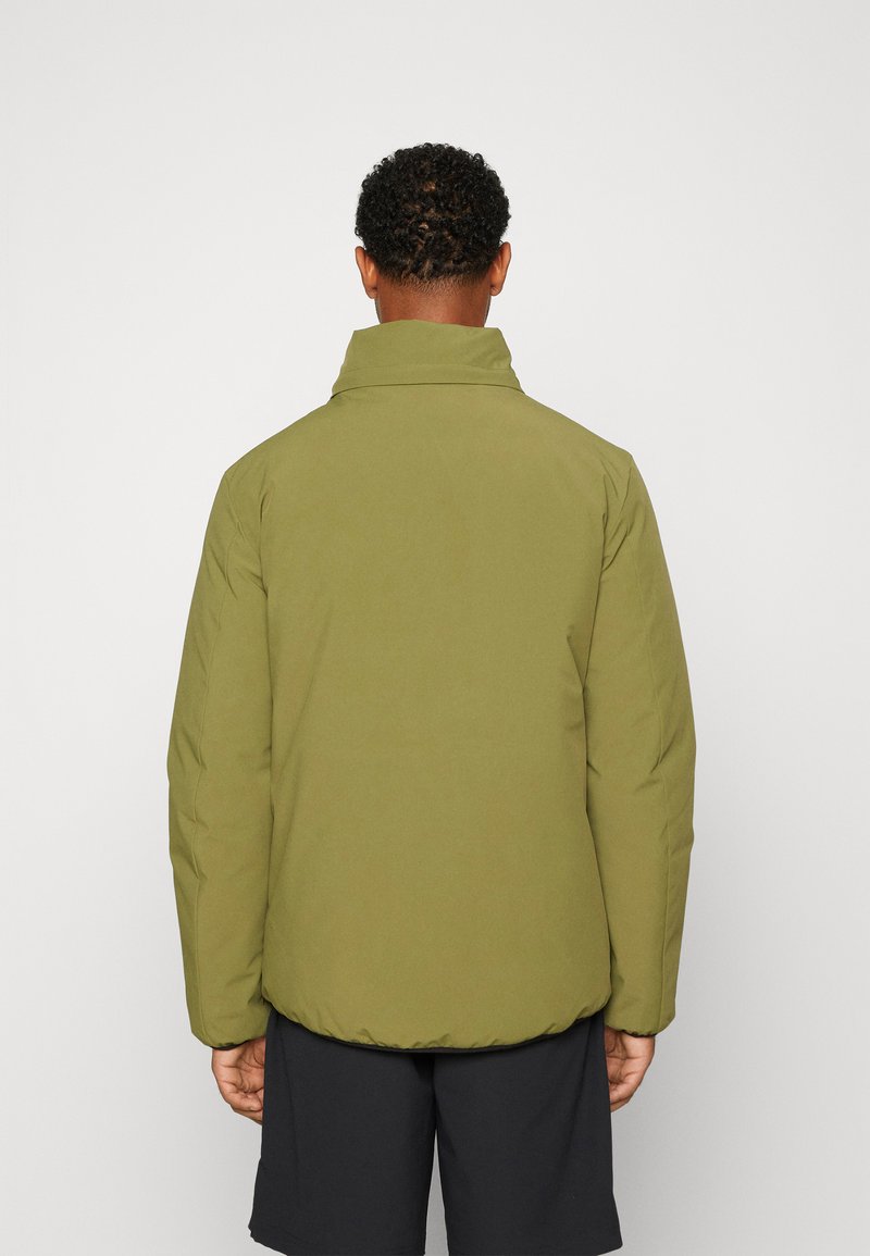 Hombre de pie de espaldas, vistiendo una chaqueta acolchada verde oliva con cuello alto y pantalones cortos negros, contra un fondo liso.