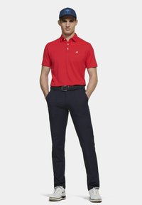 Rode polo met kraag, marineblauwe broek en witte golfschoenen. Bevat een klein logo op de borst en een blauwe pet met logo.