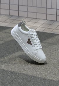 White low-top sneaker with gray heel tab, white laces, and a triangular logo on the side, placed on asphalt near tiled wall background.