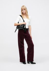 Λευκό εφαρμοστό τοπ, μπορντό παντελόνι με φαρδιά σκέλη από κοτλέ, μαύρη τσάντα με λαμπερή υφή και μαύρες μπότες με ψηλό τακούνι. Το μοντέλο στέκεται όρθιο.