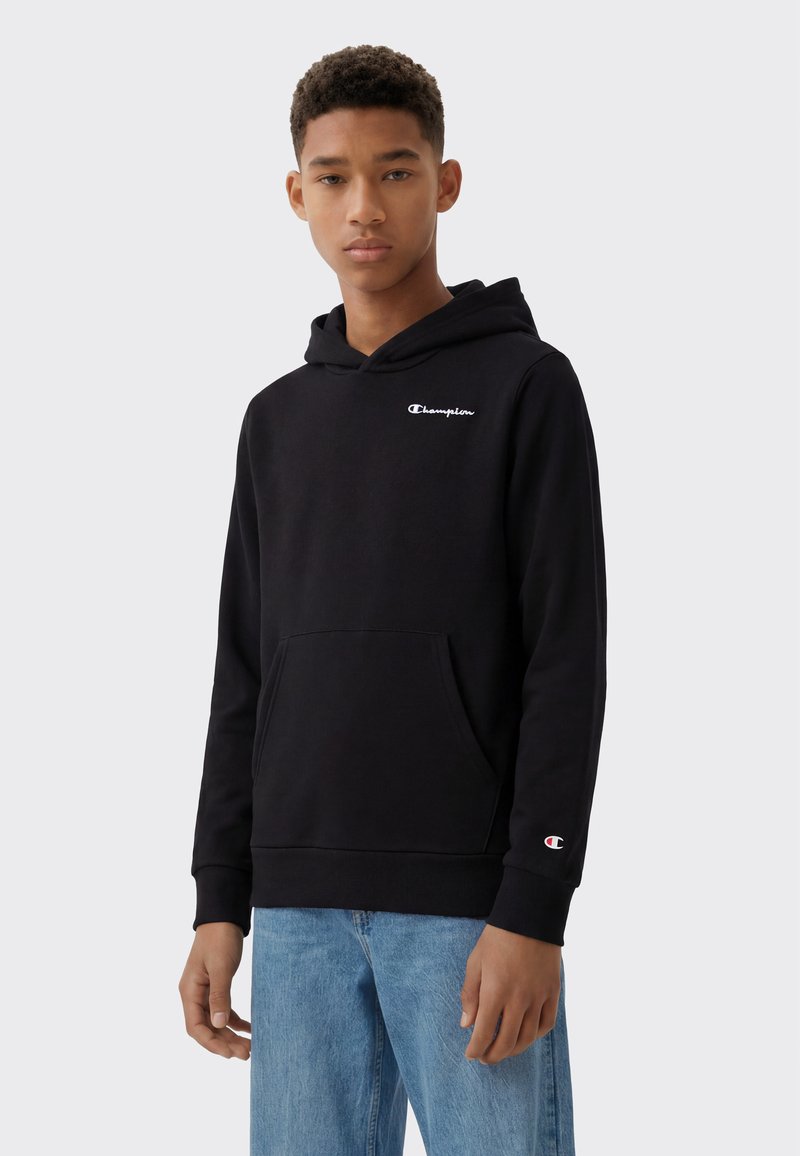 Joven con sudadera negra Champion con logo en el pecho y la manga, combinada con vaqueros azul claro, de pie frente a un fondo liso.
