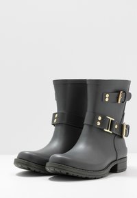 Des bottes de cheville en caoutchouc noir présentent une finition brillante, des accents en matériel doré, une sangle ajustable avec boucle et une semelle texturée noire.
