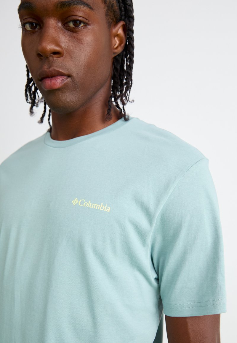 Chemise légère bleu clair à manches courtes en tissu doux. Présente un petit logo doré Columbia sur la poitrine gauche. Texture lisse, design simple.