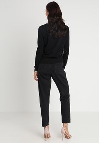 Czarny sweter z prążkowanym dołem, zestawiony z luźnymi, czarnymi jeansami z wysokim stanem. Stylizację dopełniają sandały na przeźroczystym obcasie. Gładkie tło.