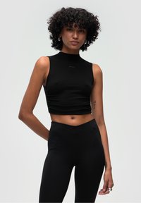 Černý bezrukávový crop top s vysokým výstřihem, vyrobený z hladkého, elastického materiálu. S kombinací s černými legínami s vysokým pasem.
