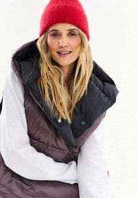 Blondínka so zapletenou červenou čiapkou, bielym tričkom s dlhým rukávom a tmavou nafúknutou vestou s veľkým golierom, usmievajúca sa na bielom pozadí.