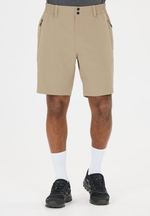 Beige shorts til knæet med elastisk linning og lynlåslommer båret med hvide mellemhøje sokker og sorte sportssko på en stående person.