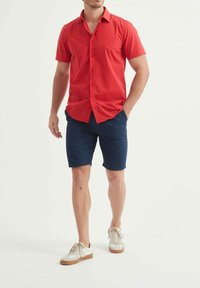Chemise rouge à manches courtes avec un col, associée à un short bleu marine et des baskets blanches avec des accents beiges.
