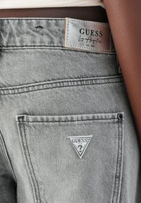 Hallitatud hall jeans kangas, millel on nahktag, kus on "GUESS" bränding ja kolmnurkne logo tagakotis. Tekstureeritud kangas näitab kerget kulumist.