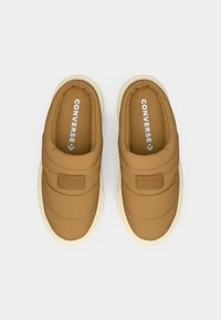 Καφέ πάνινα παπούτσια τύπου slip-on με υφή καπιτονέ, λευκές λάστιχες σόλες και εμφανές λογότυπο Converse στην εσωτερική σόλα. Λείο φινίρισμα, μινιμαλιστικός σχεδιασμός.