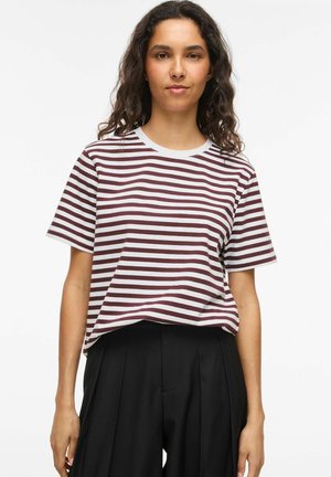 Junge Frau mit lockigem Haar, die ein weißes und dunkelrotes gestreiftes Kurzarmshirt und schwarze hoch taillierte Hose trägt, steht vor weißem Hintergrund.