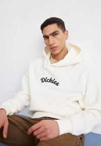 Sudadera cremosa con el logotipo bordado "Dickies" en negro, que cuenta con capucha con cordones, puños acanalados y una textura suave. Modelo sentado sobre una superficie azul.